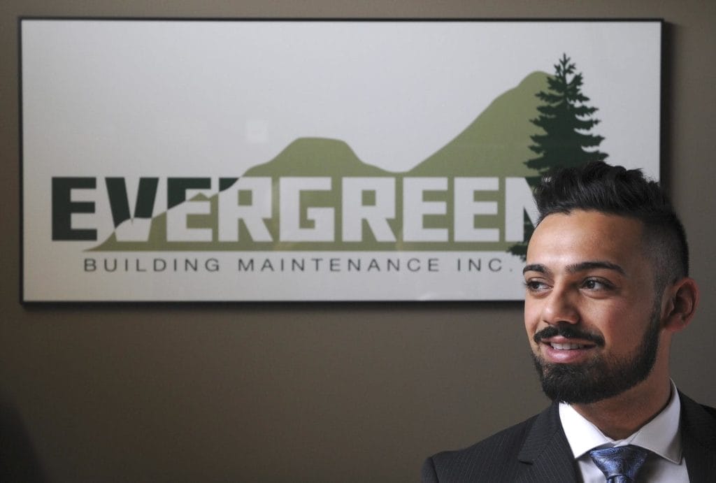A smiling man in a suit sits in front of an "EVERGREEN Building Maintenance Inc." sign featuring green mountains and a tree, representing the company’s commitment to quality commercial cleaning and property maintenance.