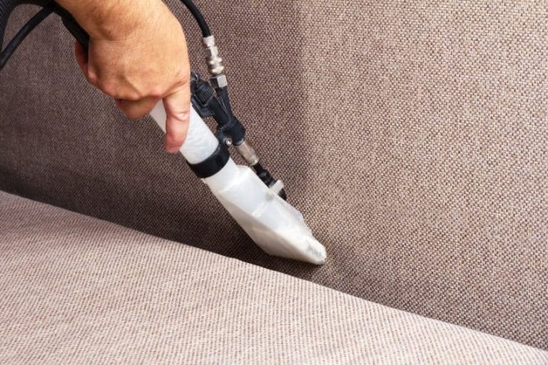 A person uses a handheld upholstery cleaning machine to clean a beige fabric sofa, focusing on the crevice between the seat and the backrest as part of thorough property maintenance.