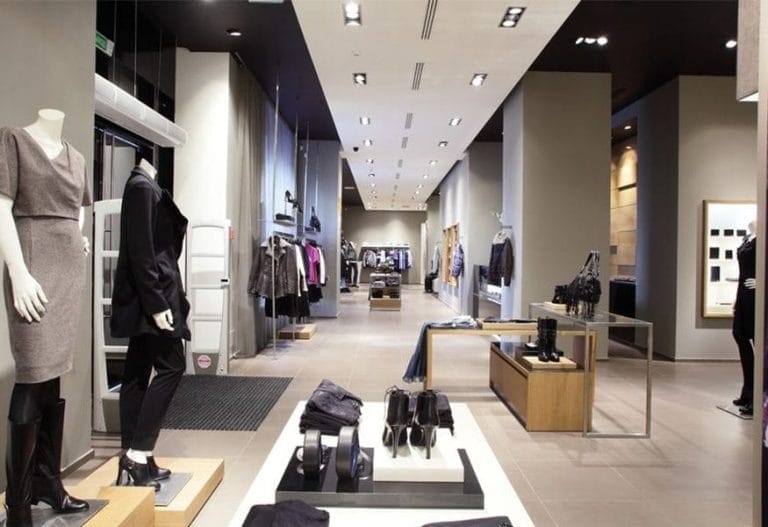 A modern clothing store interior, spotless from expert janitorial cleaning, with mannequins dressed in stylish outfits, folded clothes, and shoes displayed on tables, and racks of garments lining the walls under bright ceiling lights.