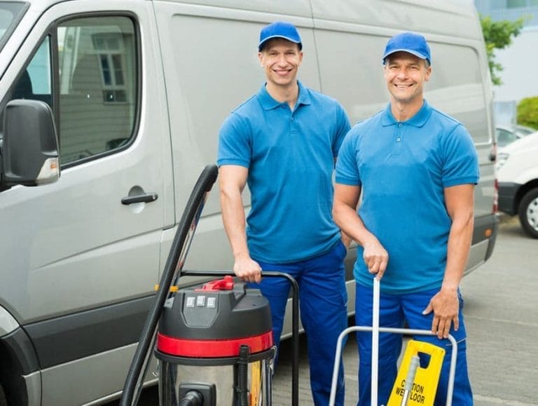 Two smiling workers in blue uniforms and caps stand in front of a van, holding cleaning equipment like a vacuum and wet floor sign, showcasing their expertise in commercial cleaning.