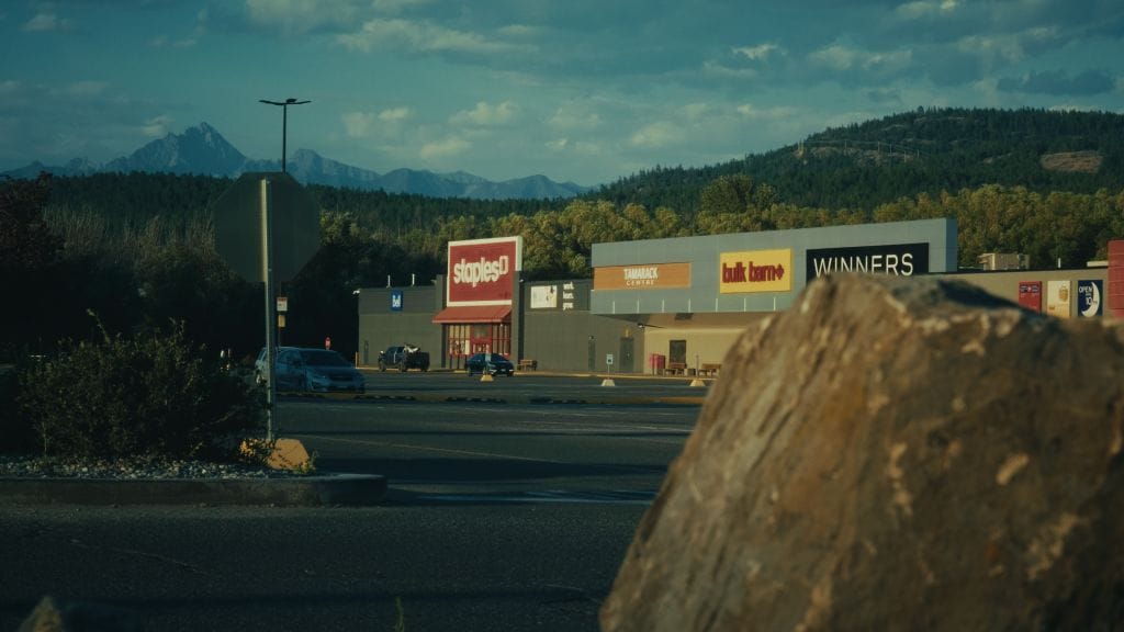 A shopping center parking lot with a few cars is seen at sunset. Stores like Simons, Bulk Barn, and Winners are visible, with trees and mountains in the background and a large rock in the foreground, reflecting well-maintained property maintenance.