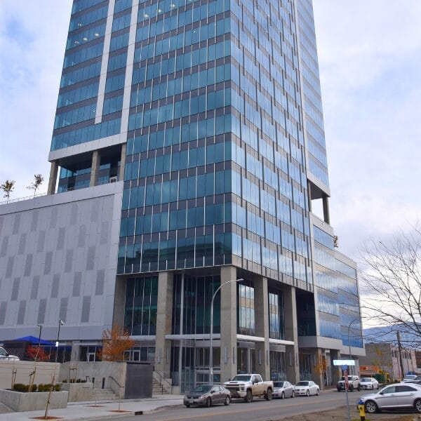 A tall, modern glass office building with blue-tinted windows stands by a city street lined with parked cars. Well-kept grounds suggest reliable property maintenance, even on this overcast day with a few leafless trees nearby.