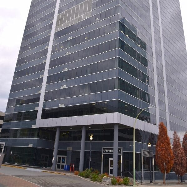 A tall, modern office building with reflective glass windows and gray trim stands on a city street, surrounded by sidewalks and a few reddish-brown trees—an ideal site for commercial cleaning services to maintain its pristine appearance.