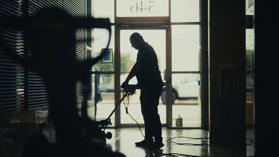 A person is silhouetted while performing janitorial cleaning on a shiny floor with a corded machine inside a building, as natural light streams through glass doors and windows in the background.