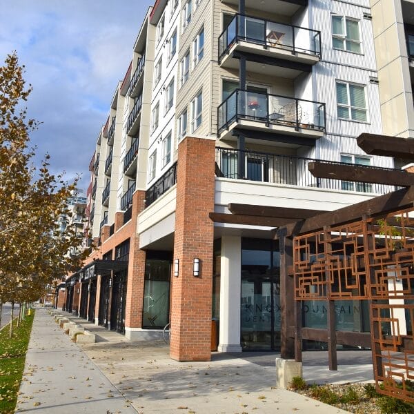 Modern apartment building with multiple balconies above ground-floor shops and businesses, brick and wood accents, a sidewalk lined with young trees, and a clear sky—ideal for those seeking professional property maintenance and janitorial cleaning services.