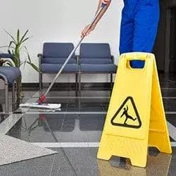 A person mops the shiny tile floor of a waiting area with chairs and plants, while a yellow caution sign warns of a wet floor—highlighting the importance of commercial cleaning and property maintenance.