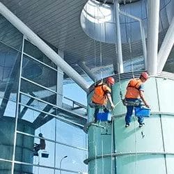 Two workers, equipped with safety harnesses and helmets, perform commercial cleaning on a modern glass building while suspended by ropes. Each holds a blue bucket, with their reflections visible on the shimmering glass behind them.
