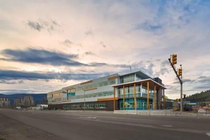 A modern, multi-story building with large glass windows and yellow accents stands at a street corner under a cloudy sky, highlighting the importance of property maintenance for such structures amid busy streets and distant city buildings.