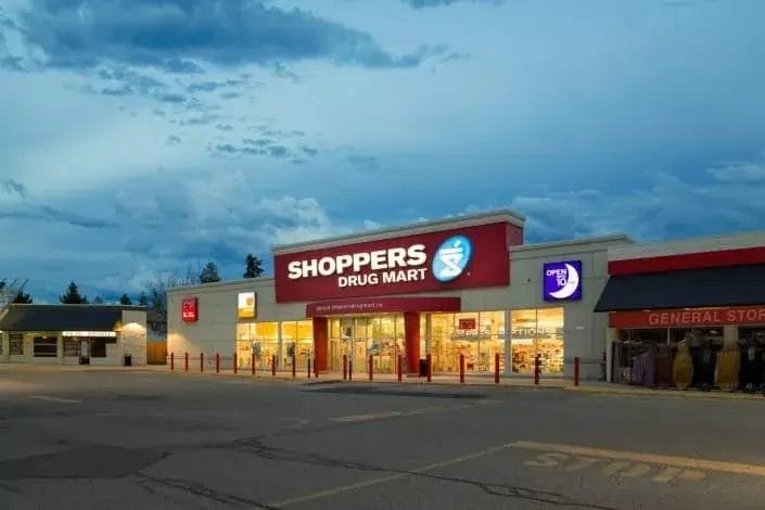 A Shoppers Drug Mart store is brightly lit at dusk, its red and white sign glowing above the entrance. An illuminated "open 24 hours" sign welcomes customers, while the well-kept parking lot highlights reliable property maintenance.