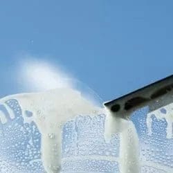 A close-up of a squeegee cleaning soapy water off a glass window, leaving a clear streak against a blue background—perfect for showcasing professional Commercial Cleaning results.