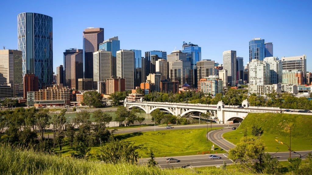Downtown Calgary skyline with modern skyscrapers, a white bridge over the Bow River, green park areas, and well-maintained properties benefiting from expert commercial cleaning under a clear blue sky on a sunny day.