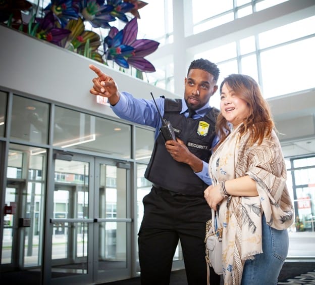 A security guard in uniform points while speaking to a smiling woman in a lobby with large windows and colorful flower decorations overhead. The guard holds a walkie-talkie.