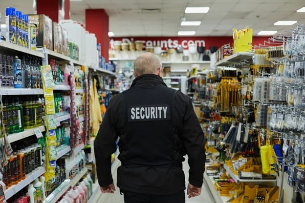 A security guard wearing a black jacket with "SECURITY" on the back walks down an aisle in a hardware store, ensuring client-centric security among rows of tools and supplies.