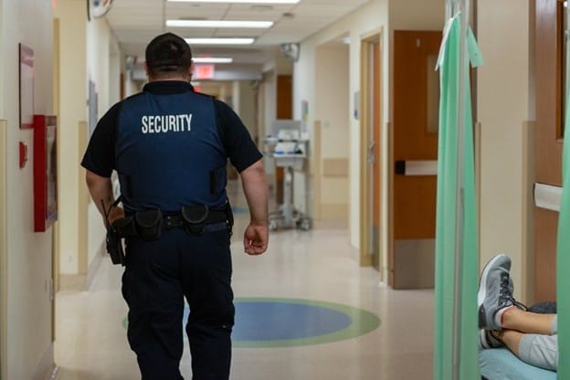 A security guard in uniform, embodying client-centric security, walks down a brightly lit hospital hallway, with a person’s legs visible on a bed to the right and hospital rooms lining both sides.