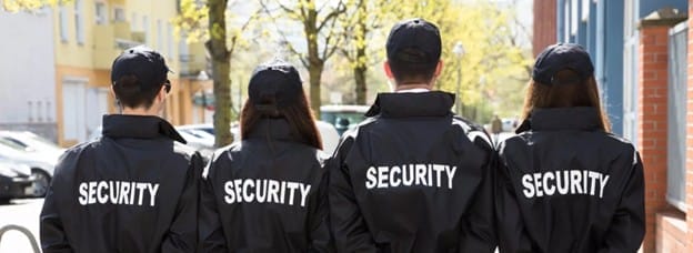 Four people in black jackets and caps labeled "SECURITY" stand side by side outdoors, embodying a client-centric security approach on a sunny day, with trees and buildings visible in the background.