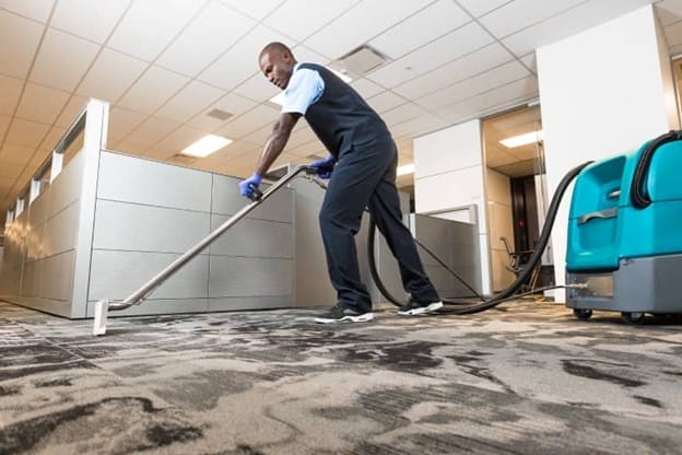 A person in uniform uses commercial carpet cleaning equipment to clean the floor of an office space with cubicles and overhead lighting.