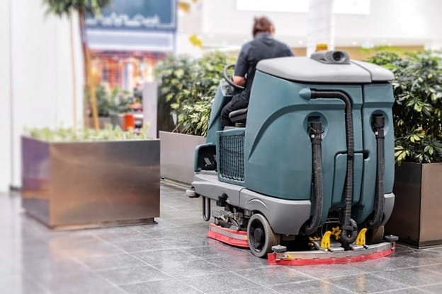 A person operates a large industrial floor cleaning machine in an indoor area with tile flooring and potted plants.
