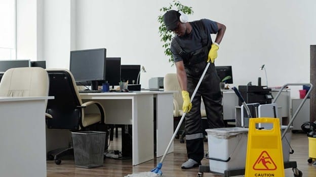 A person wearing headphones, gloves, and work overalls is mopping the floor in an office with desks and computers. A yellow "Caution: Wet Floor" sign is placed nearby. Cleaning equipment is visible.