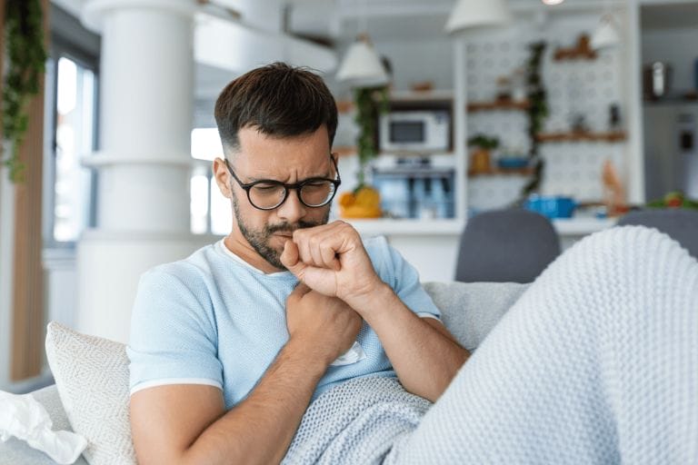 A man with glasses sits on a couch, wrapped in a blanket and coughing into his fist, appearing unwell. The bright, modern living room highlights the importance of regular commercial cleaning for a healthier environment.