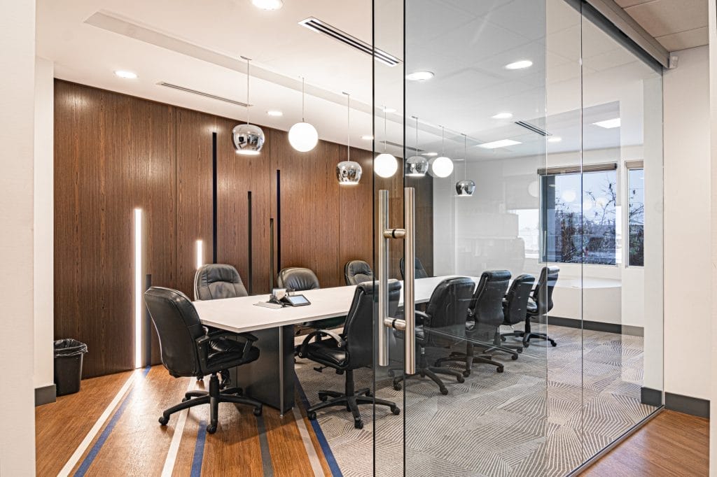 A modern conference room with a long white table, black chairs, pendant lights, wooden wall panels, and floor-to-ceiling glass walls; outside, a window shows trees and buildings.
