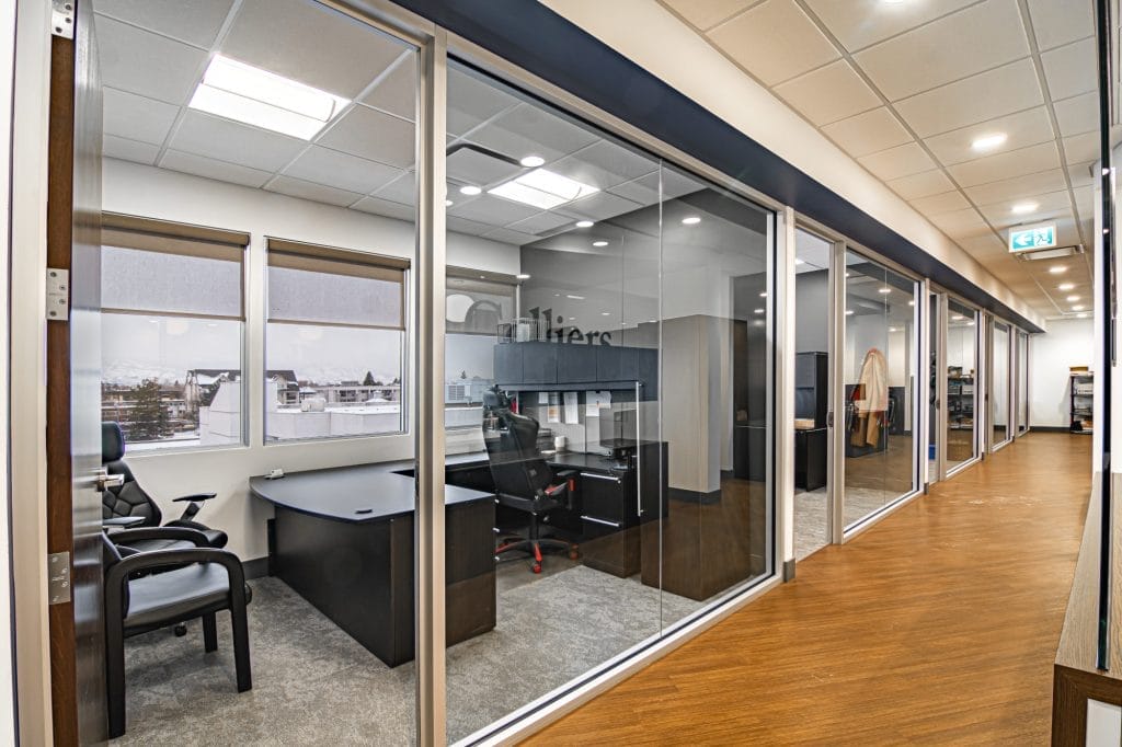Modern office hallway with glass-walled private offices, black desks and chairs, natural light from large windows, wood flooring, and a view of buildings outside. Ceiling lights provide bright indoor lighting.
