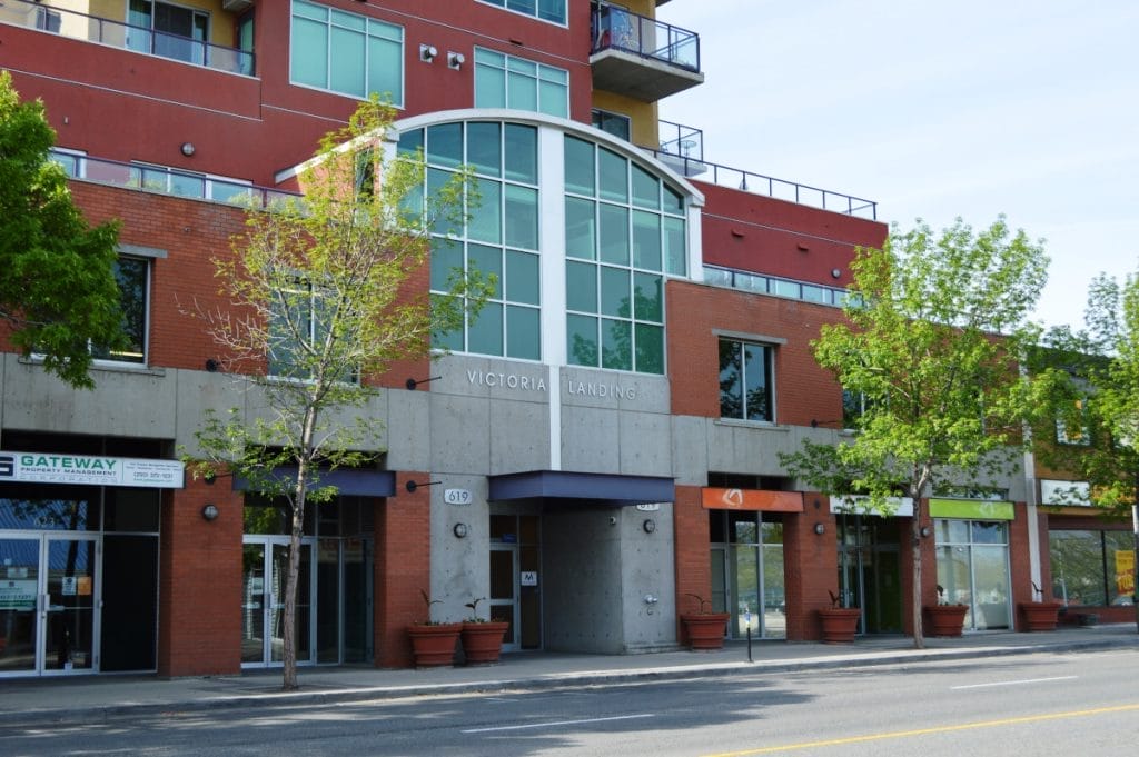 A modern red-brick and concrete building with large windows, labeled "Victoria Landing," sits at the Evergreen Business Centre. Gateway Realty and other businesses are on the ground floor, while trees line the sidewalk in front.