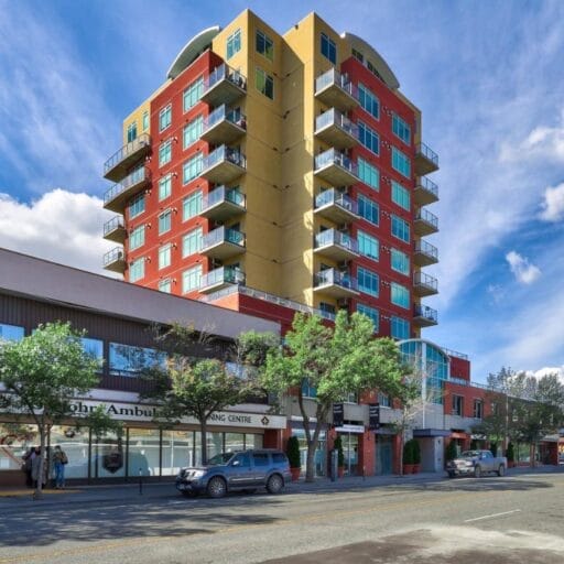 A colorful multi-story apartment building with yellow and red walls rises above Evergreen Business Centre and nearby shops and trees on a sunny urban street, with cars parked along the sidewalk. Blue sky and clouds fill the background.