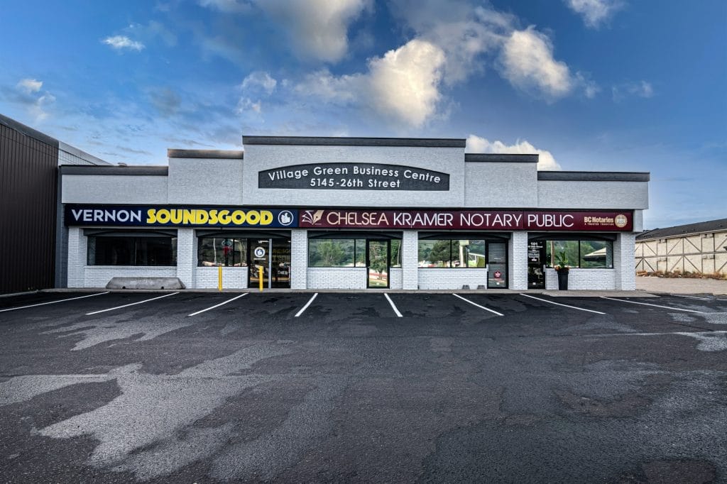 Village Green Business Centre is a one-story strip mall featuring signs for Vernon Soundsgood, Chelsea Kramer Notary Public, and RE/MAX offices, with an empty parking lot in front beneath a partly cloudy sky.