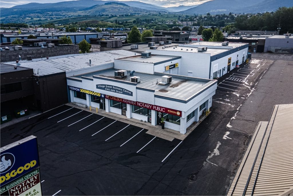 The Village Green Business Centre, home to businesses including a notary public, is surrounded by an empty parking lot and set against a backdrop of a hilly, green landscape beneath a partly cloudy sky.
