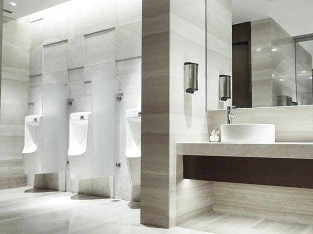 A modern public restroom featuring three urinals with frosted glass dividers, a large mirror, two soap dispensers, and a countertop with a vessel sink in a sleek, neutral-toned design.