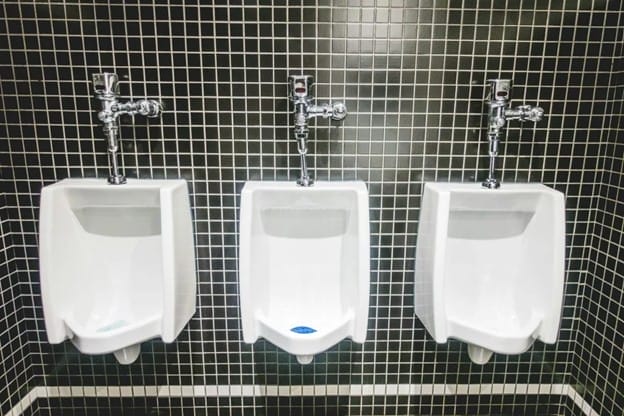 Three white porcelain urinals are mounted on a black tiled wall in a public restroom. Each urinal has a chrome flush valve above it, evenly spaced across the wall.