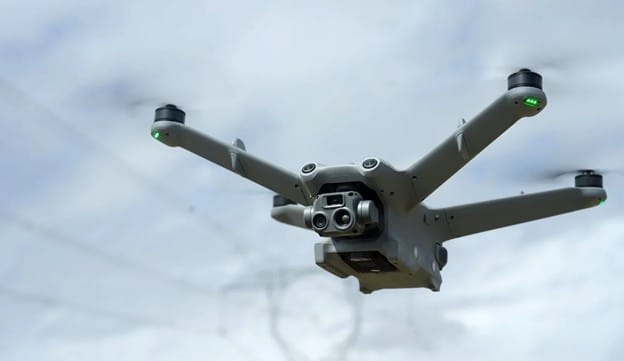 A close-up of a gray quadcopter drone with a camera, flying outdoors against a cloudy sky. Green indicator lights are visible on its arms.