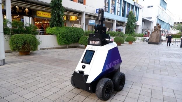 A wheeled security robot labeled "XAVIER" patrols an outdoor pedestrian area, surrounded by plants, shops, and buildings. The robot features cameras and sensors on top.