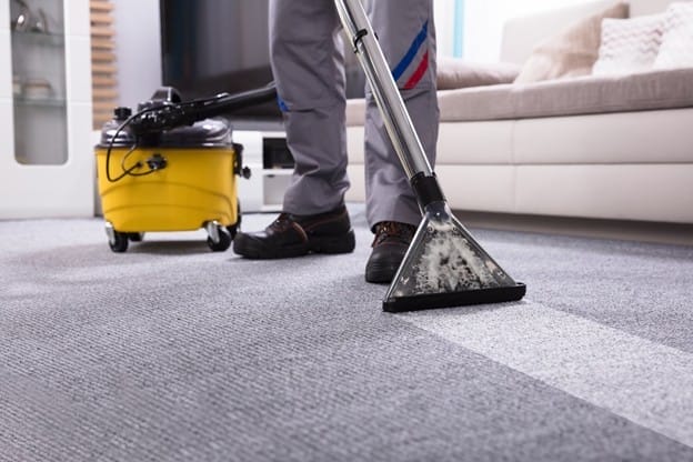 A person uses a carpet cleaning machine to deep clean a gray carpet in a living room, with a yellow vacuum unit behind them and a sofa in the background.