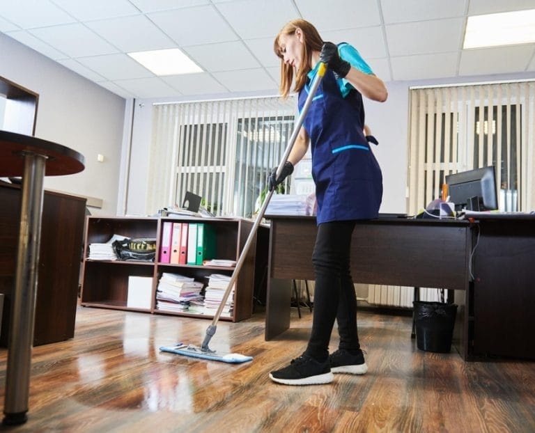 A woman in a blue uniform and gloves is mopping the floor in an office with desks, computers, office chairs, and shelves filled with files. The room is well-lit with large windows and vertical blinds.