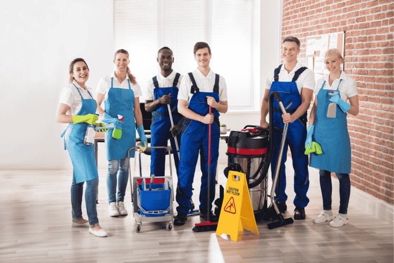 A group of six smiling janitors in blue uniforms stand together with cleaning supplies, including a mop, bucket, vacuum, and caution sign, in a bright, modern room with wooden floors and brick walls.