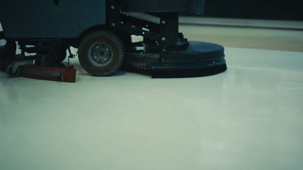 A close-up of an industrial floor cleaning machine from Evergreen Building Maintenance shows rotating brushes scrubbing a smooth, light-colored floor. Only the lower part of the machine and its wheels are visible.