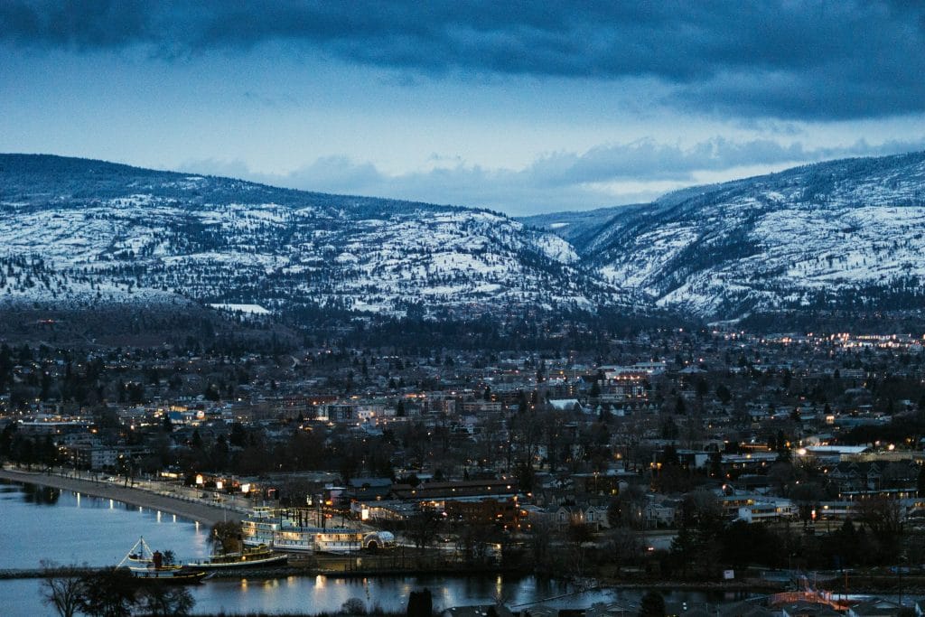 Cityscape at dusk with snow-dusted mountains in the background, a waterfront dotted with docks and buildings—some maintained by Evergreen Building Maintenance—and scattered lights illuminating the town under a cloudy sky.