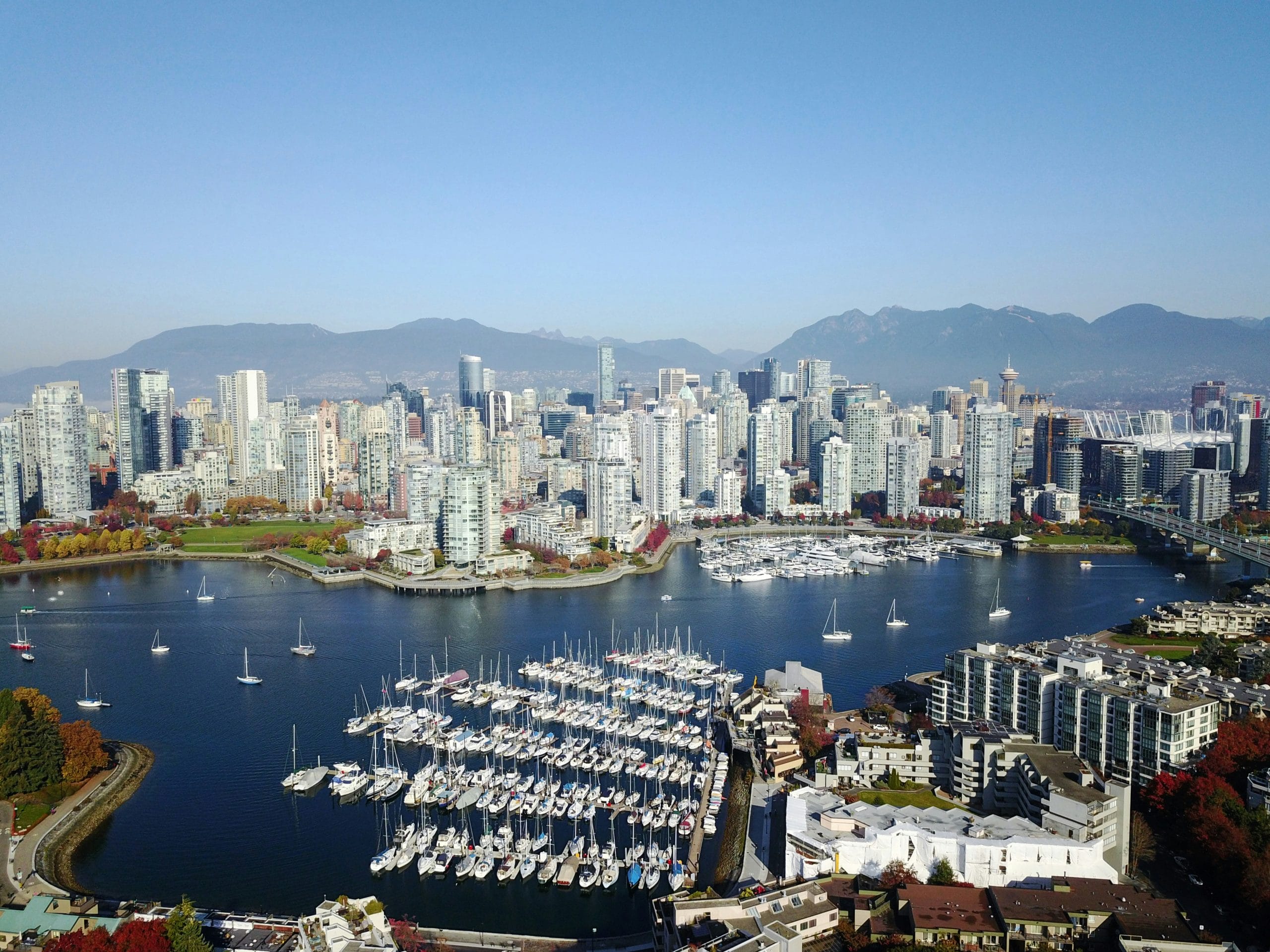Aerial view of a marina filled with boats in front of a city skyline, including the Evergreen Building Maintenance tower, surrounded by water and mountains under a clear blue sky.