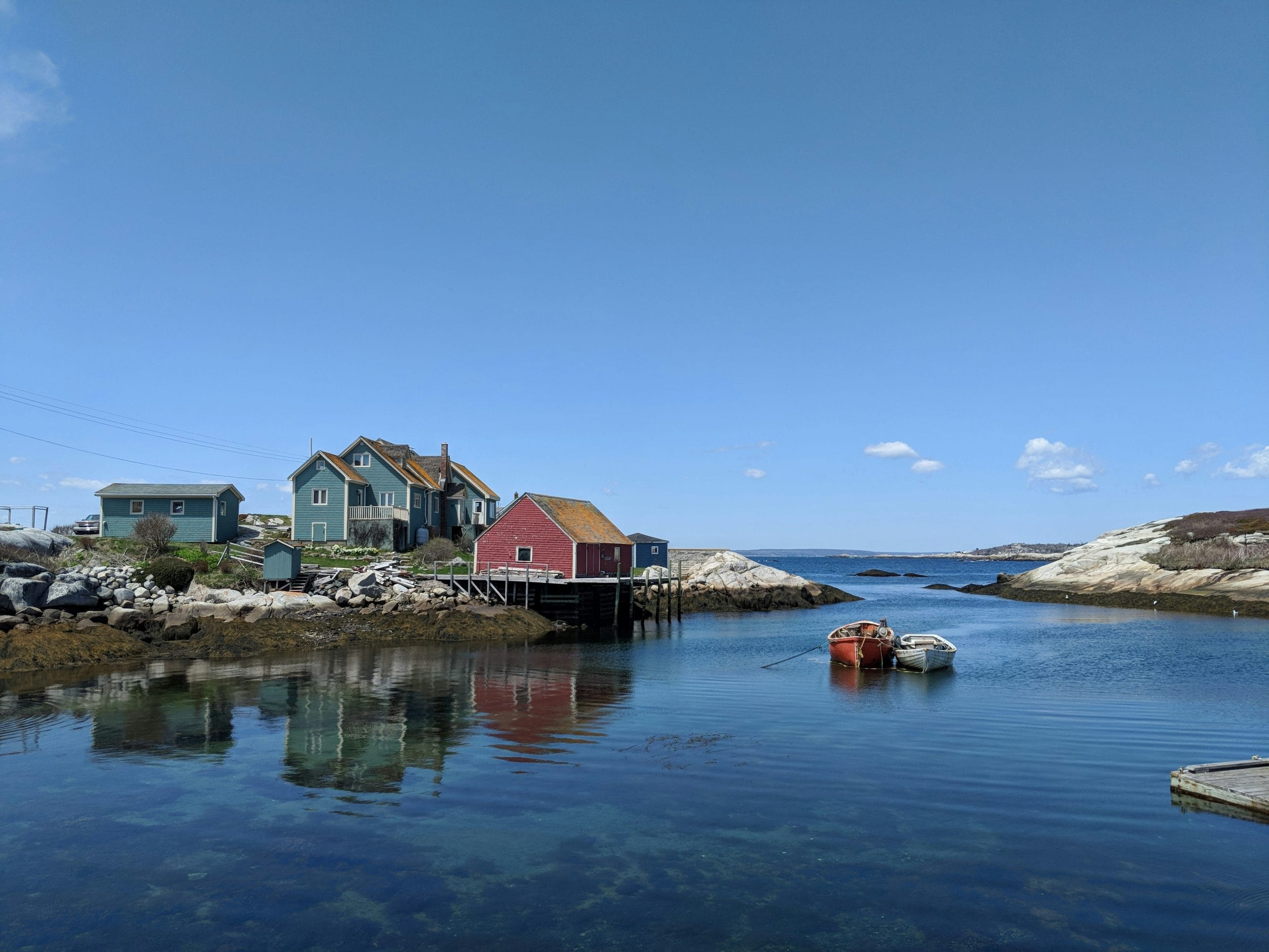 Coastal scene with colorful wooden houses, including a red one on stilts by the water, rocky shoreline maintained by Evergreen Building Maintenance, two small boats in a calm bay, and a clear blue sky.