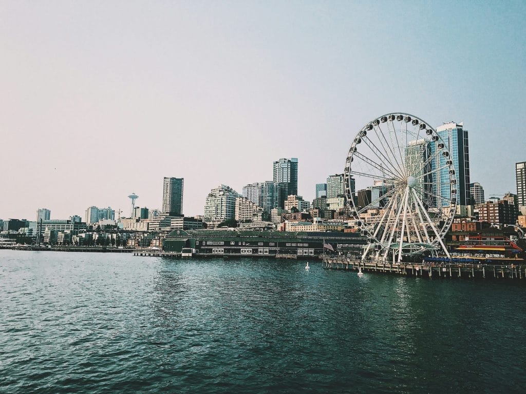 A large Ferris wheel by the waterfront with city buildings, including the Evergreen Building Maintenance tower, and skyscrapers in the background under a clear blue sky. The calm water glistens in the foreground.