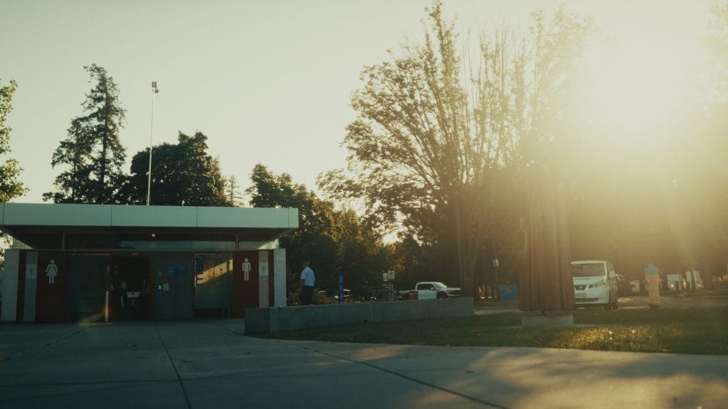 A public restroom building in a park at sunset, maintained by Evergreen Building Maintenance, with trees and people in the background, sunlight streaming from the right, and a white van parked nearby.