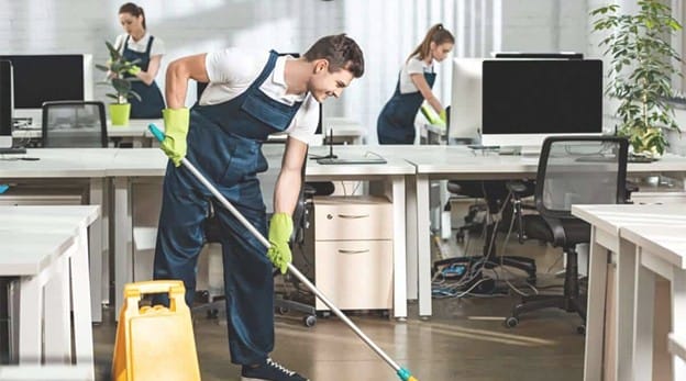 Three people in uniforms and gloves are cleaning a modern office; one mops the floor near a wet floor sign while two others clean desks and surfaces with computers in the background.