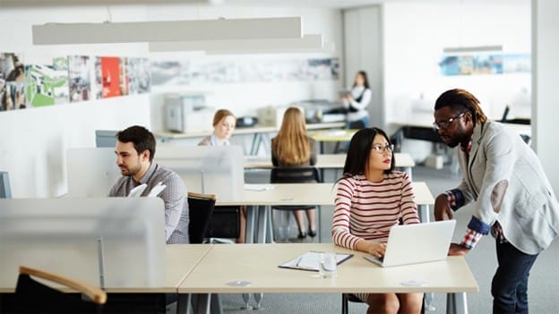 Several people work at desks in a modern open-plan office. Two colleagues are talking at one desk, while others work independently at their computers. The atmosphere appears collaborative and professional.