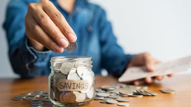 A person places a coin into a glass jar labeled "SAVINGS," surrounded by scattered coins on a wooden table. The person is wearing a blue denim shirt.