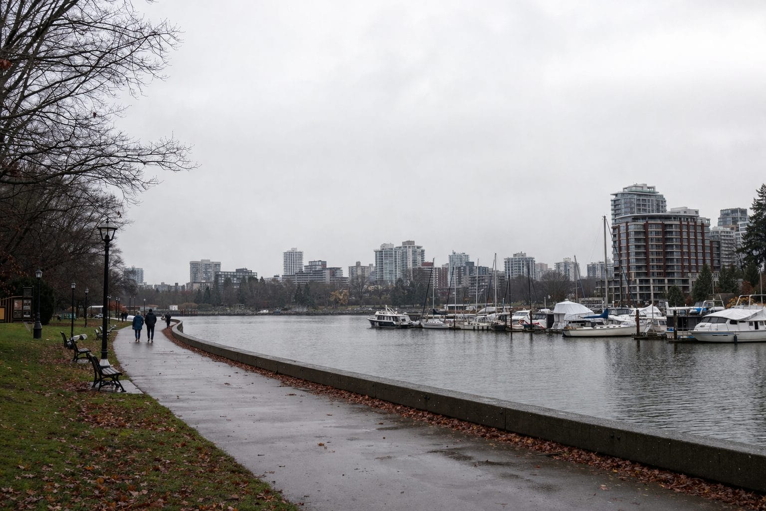 A cloudy waterfront scene shows a paved path beside a marina with docked boats, a few people walking, benches, and high-rise buildings in the background. Leafless trees line the left side of the path.
