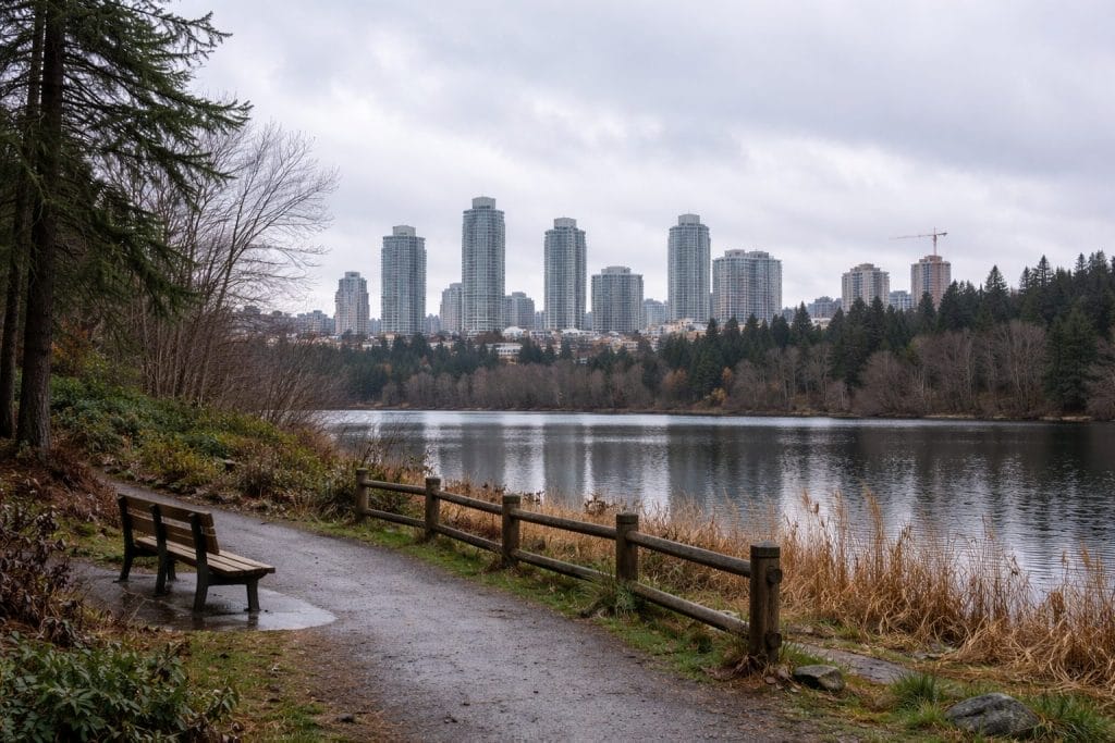 A park path with a bench overlooks a calm lake bordered by trees, with tall city buildings and cloudy skies in the background.