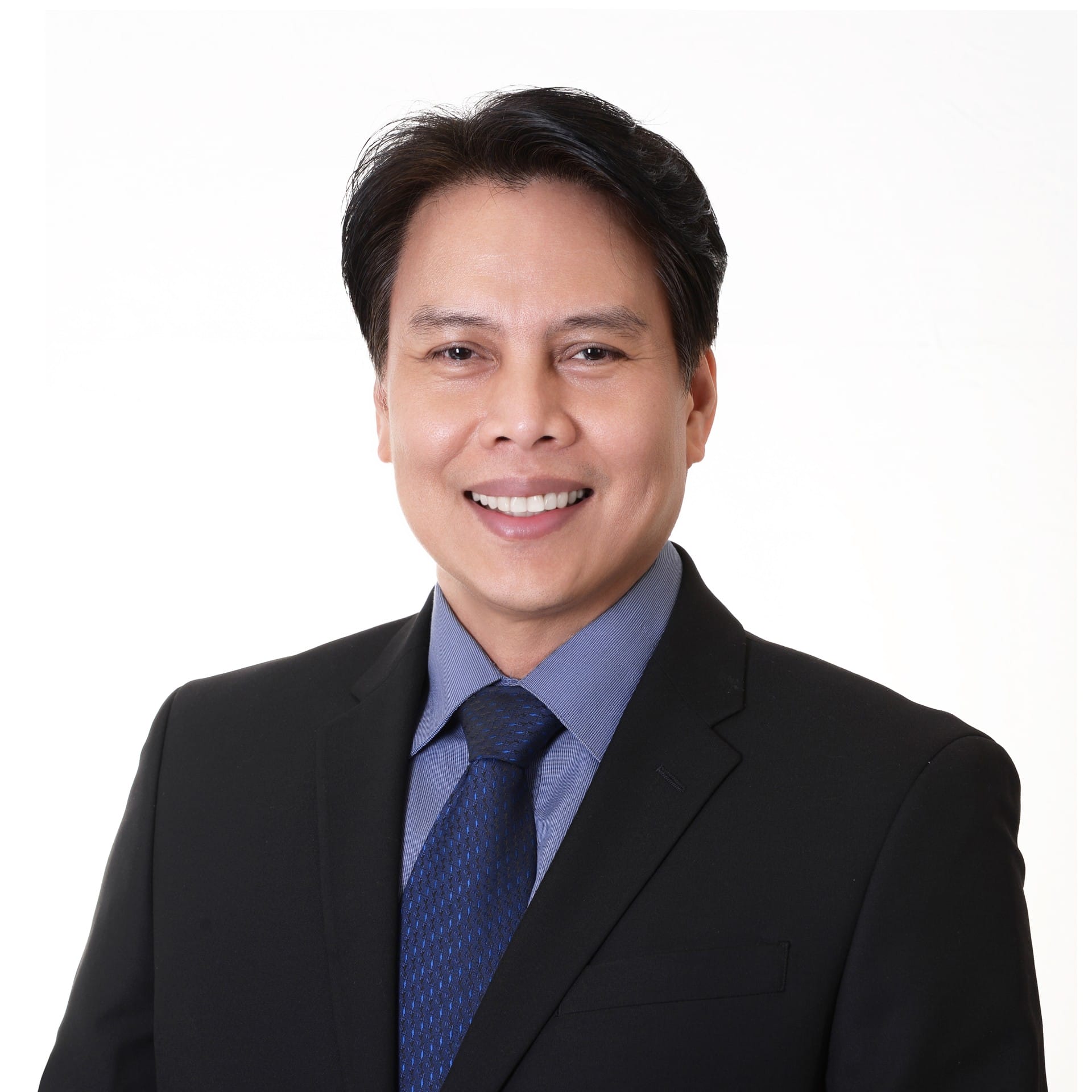 A man with short black hair smiles while wearing a black suit, blue dress shirt, and blue patterned tie, representing the Evergreen team against a plain white background.