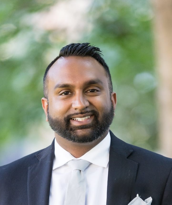 A man with short dark hair and a beard, wearing a black suit, white shirt, and light gray tie, smiles outdoors with a blurred green background, representing the professionalism of the Evergreen team.